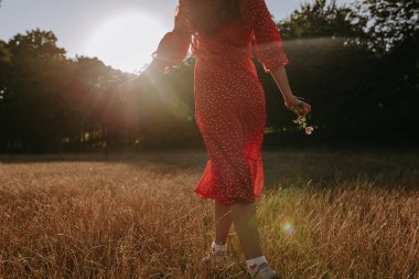 Arkaya bakan kadın portresi. Yazın kısa kırmızı elbiseli. Gün batımında çayırların arasından ormana doğru yürüyor.