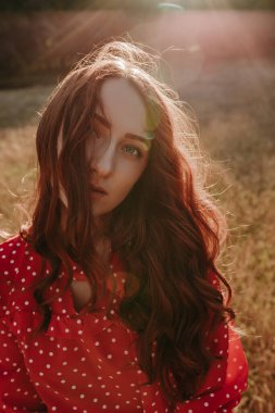 Kızıl saçlı kadın, ormanın arka planındaki kuru çim tarlasında kamera önünde poz veriyor.