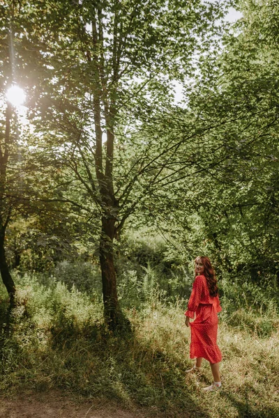 穿着红衣服的年轻漂亮的红头发女人手牵着花走过草地 森林荒地上笑容满面的女性的低视角画像 — 图库照片