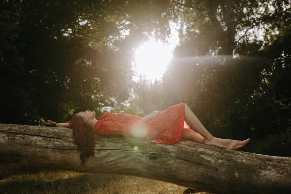 一个身穿红色夏装的年轻貌美的女人 白斑斑斑地躺在森林中央一棵倒下的干树干上 — 图库照片