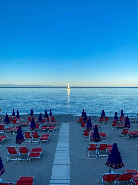 Sahilin önündeki körfezde demirlemiş bir yelkenli üzerinde şemsiyeler ve straio, Noli, Liguria - İtalya