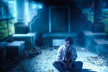 Mezarlıkta oturan, üzerinde kan ve yara olan korkunç bir zombi.