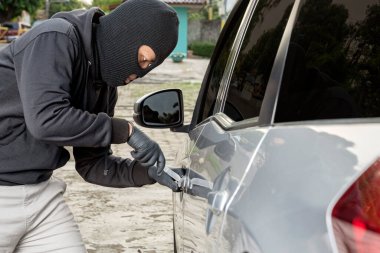 Araba hırsızı, elinde bir aletle sokakta araba kırmaya çalışıyor.
