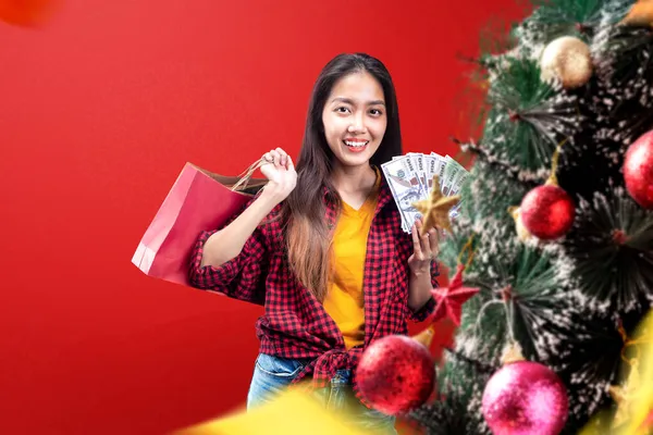 アジアの女性がお金とショッピングバッグを持っている色の背景にクリスマスツリーを飾る ボクシングの日 — ストック写真