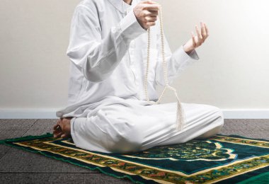 Müslüman bir adam ellerinde dua boncuklarıyla dua ederken beyaz duvar arkaplanlı bir halının üzerinde oturuyor.