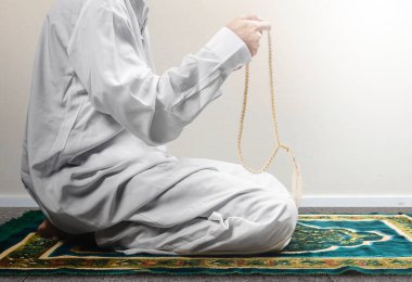 Müslüman bir adam ellerinde dua boncuklarıyla dua ederken beyaz duvar arkaplanlı bir halının üzerinde oturuyor.