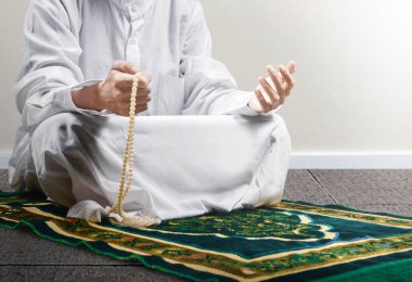 Müslüman bir adam ellerinde dua boncuklarıyla dua ederken beyaz duvar arkaplanlı bir halının üzerinde oturuyor.