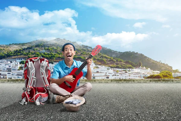 Asian man with a beanie hat and a backpack playing guitar to make money on the street with blue sky background