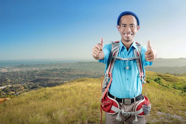 An Asian man with a beanie hat and a backpack showing a thumb up gesture standing on the hill with a landscape view background