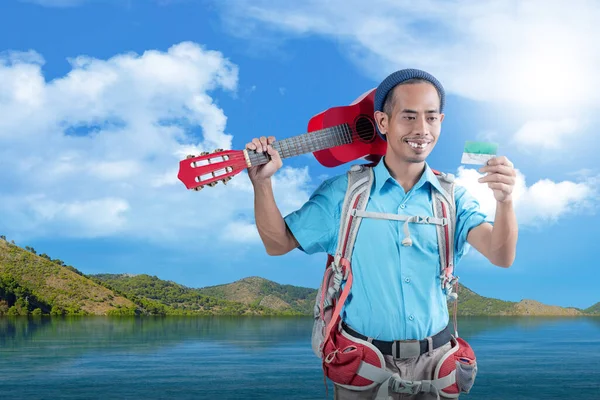 Asian man with a beanie hat and a backpack holding card and carrying a guitar standing with a lake and hill view background