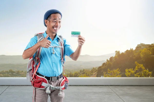 Asian man with a beanie hat and a backpack holding card with a thumb up gesture standing on the street with a landscape view background