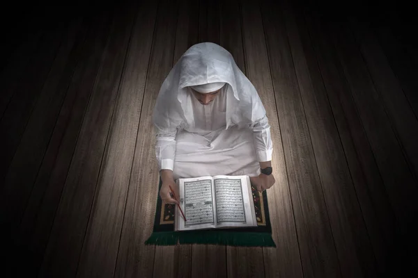 Arab Muslim man sitting and reading the Quran on the prayer rug with a wooden floor background