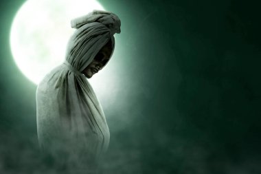 Pocong is covered with a white linen shroud standing with full moon background. Halloween concept