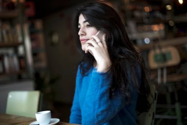 Cep telefonuyla konuşan mutlu bir kadın. Kafede oturuyor. Camdan görülüyor.