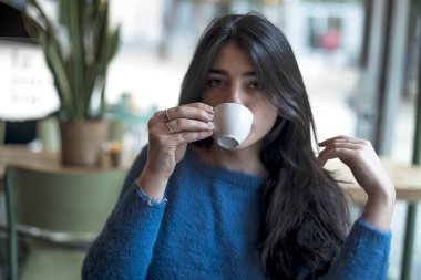 Genç model uzun siyah saçlı kadın brunch sırasında bir kahve dükkanında oturuyor. Latin kadın kafede masada otururken aşağı bakıyor..