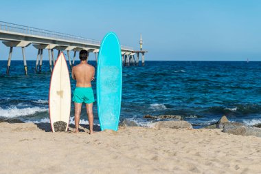 Deniz kıyısındaki sörf yerinde sörf tahtası olan yakışıklı bir adama yaklaş ve dalgayı bekle. Arkadan görüntüle.