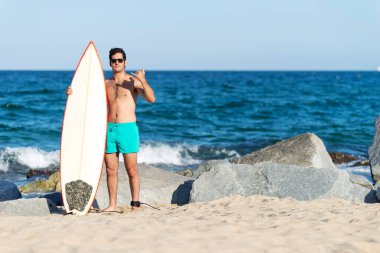 Güneş gözlüklü İspanyol sörfçü deniz kıyısında sörf tahtası tutuyor.