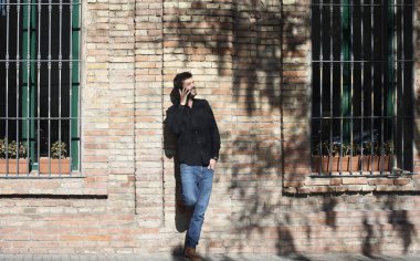 Güneşli bir günde duvara yaslanırken cep telefonuyla konuşan kendine güvenen bir adam.