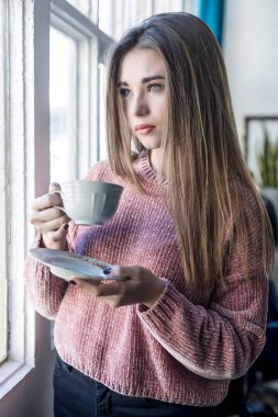 Elinde bir fincan çayla pencerenin yanında duran genç bir kadın..