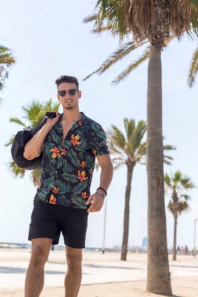 Young man walking on city wearing casual clothes and sunglasses with shorts flowered shirt holding travel bag