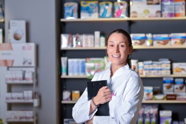 İlaç raflarının yanında duran genç eczacı elinde not defteri tutuyor.