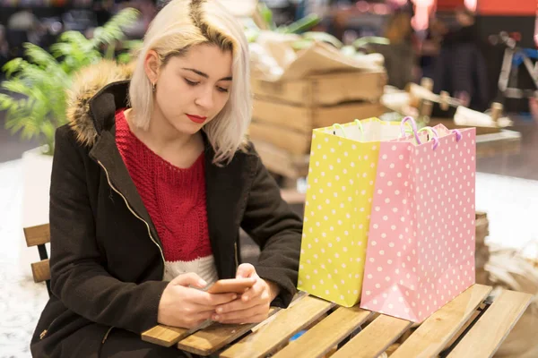 美丽快乐的女人的画像 她坐在购物中心里 提着五颜六色的购物袋 用智能手机应用程序 看屏幕 发短信 — 图库照片