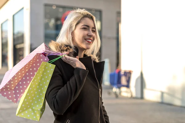 年轻迷人的金发女人穿着黑色夹克站在城市街道的背景下 拿着很多购物袋 — 图库照片