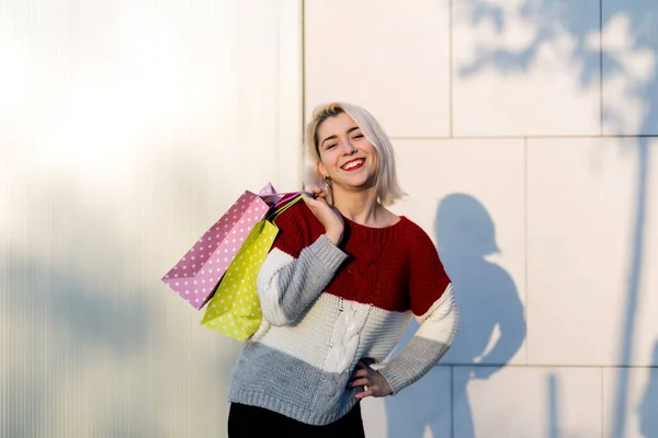 年轻快乐微笑的女人站在白墙旁边笑着拿着购物袋看着相机的画像 — 图库照片
