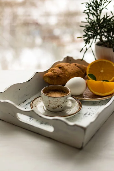 Tazza di caffè, croissant, uovo, frutta fresca arancione su vassoio di legno bianco sullo sfondo della finestra sfocata — Foto Stock