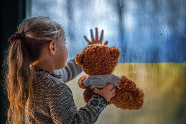 Little Girl Holds Teddy Bear Looks Window Blue Yellow Flag — Stock Photo, Image