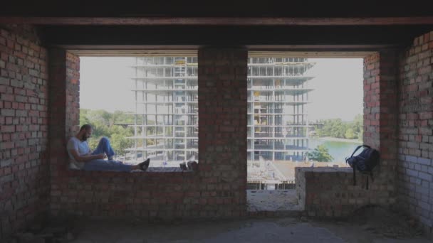 Ένας νεαρός άνδρας κάθεται στο παράθυρο στο παρασκήνιο ενός εργοταξίου και κοιτάζει το τηλέφωνο. Ένας νεαρός εργάτης σε μια οικοδομή κοιτάζει το τηλέφωνο. Ο τύπος κάθεται στο παράθυρο του σπιτιού. — Αρχείο Βίντεο