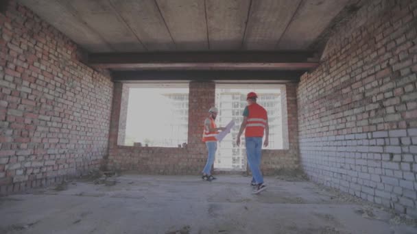 Ingenjörer framför en flervåningshus. Två ingenjörer på en byggarbetsplats tittar på ritningen. Två byggare på bakgrunden av huset — Stockvideo