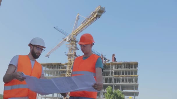 Dos ingenieros en un sitio de construcción están mirando el dibujo. Ingenieros frente a un edificio de varios pisos. Dos constructores en el fondo de la casa — Vídeo de stock