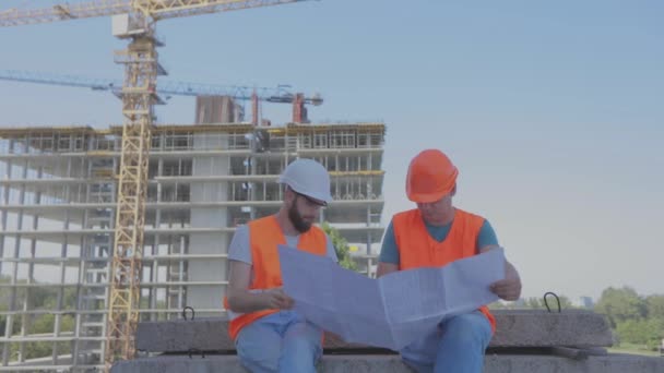 Zwei Bauarbeiter im Hintergrund des Hauses. Ingenieure vor einem mehrstöckigen Gebäude. Zwei Ingenieure auf einer Baustelle betrachten die Zeichnung. — Stockvideo