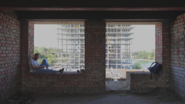 Killen sitter på fönstret i huset. En ung man sitter vid fönstret mot bakgrunden av en byggarbetsplats och tittar på telefonen. En ung man på en byggarbetare tittar på telefonen. — Stockvideo