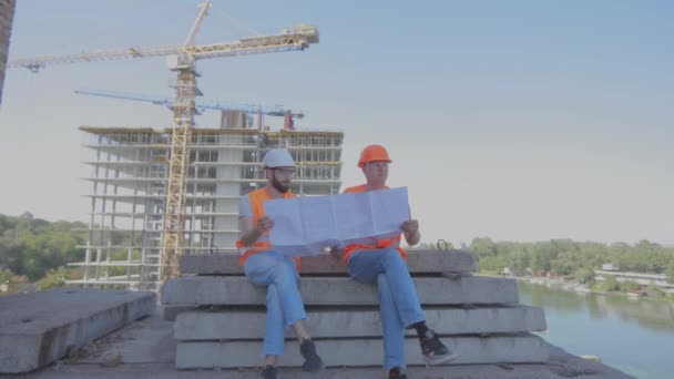 Ingenieros frente a un edificio de varios pisos. Dos ingenieros en un sitio de construcción están mirando el dibujo. Dos constructores en el fondo de la casa — Vídeo de stock