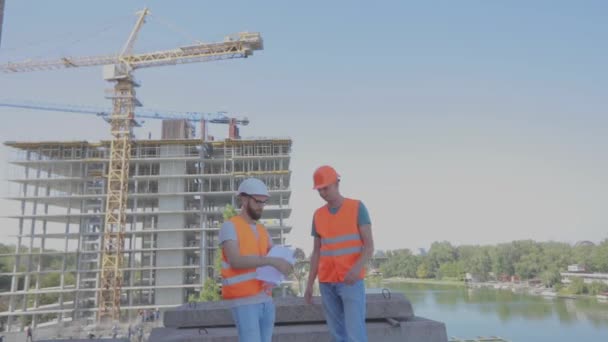 Två ingenjörer på en byggarbetsplats tittar på ritningen. Ingenjörer framför en flervåningshus. Två byggare på bakgrunden av huset — Stockvideo