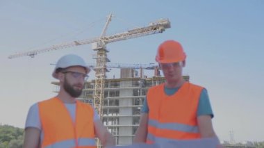 Mühendisler çok katlı bir binanın önünde. İnşaat alanındaki iki mühendis çizime bakıyor. Evin arka planında iki inşaatçı var.
