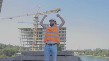 Bir mühendis inşaat alanında sevinir. Mühendis inşaat alanında neşeyi canlandırıyor. İnşaat alanında genç bir mühendis ellerini kaldırıyor.
