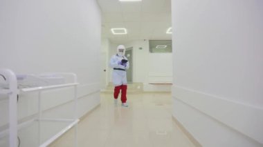 Elinde tabletle bir doktor hastane koridorunda yürüyor. Doktor modern kliniğin koridoru boyunca yürür. Koruyucu kıyafetli bir doktor.