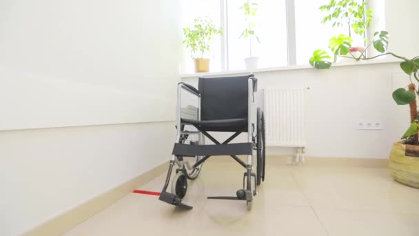 Lege rolstoel tegen de achtergrond van een helder raam. Een rolstoel voor mindervaliden in de witte hal. Lege rolstoel voor mensen met een handicap in de ziekenhuisgang. — Stockvideo