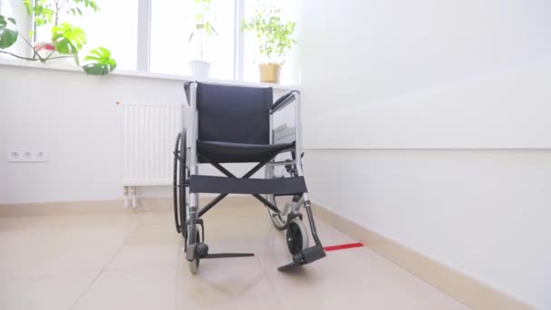 Parlak bir pencerenin arkasında boş bir tekerlekli sandalye var. Beyaz koridorda engelliler için tekerlekli sandalye. Hastane koridorunda engelliler için boş tekerlekli sandalye.. — Stok video