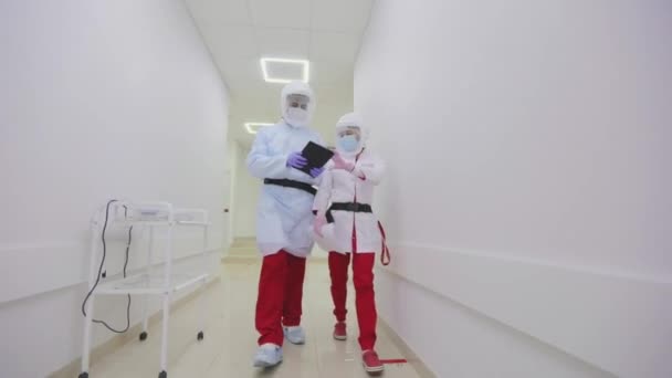 Lékaři v ochranných oblecích procházejí nemocniční chodbou. Lékaři v antivirové ochraně kráčejí světlými nemocničními chodbami — Stock video