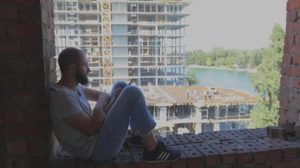 Mladý muž sedí u okna na pozadí staveniště a dívá se na telefon. Mladý muž u stavebního dělníka se podívá na telefon. Ten chlap sedí na okně domu. — Stock video