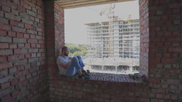 Ένας νεαρός άνδρας κάθεται στο παράθυρο στο παρασκήνιο ενός εργοταξίου και κοιτάζει το τηλέφωνο. Ένας νεαρός εργάτης σε μια οικοδομή κοιτάζει το τηλέφωνο. Ο τύπος κάθεται στο παράθυρο του σπιτιού. — Αρχείο Βίντεο