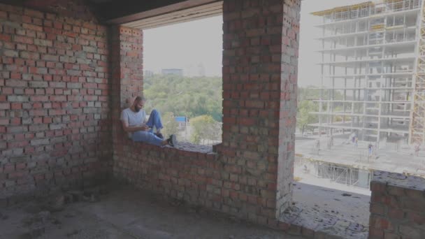 Een jongeman zit voor het raam tegen de achtergrond van een bouwplaats en kijkt naar de telefoon. Een jongeman bij een bouwvakker kijkt naar de telefoon. De man zit op het raam van het huis. — Stockvideo