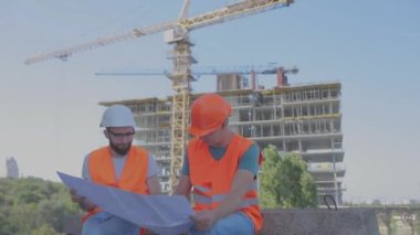 Mühendisler çok katlı bir binanın önünde. İnşaat alanındaki iki mühendis çizime bakıyor. Evin arka planında iki inşaatçı var.