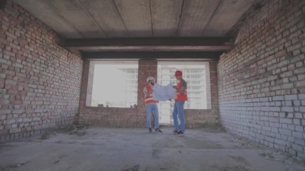 Mühendisler çok katlı bir binanın önünde. İnşaat alanındaki iki mühendis çizime bakıyor. Evin arka planında iki inşaatçı var. — Stok video