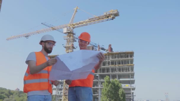 İnşaat alanındaki iki mühendis çizime bakıyor. Mühendisler çok katlı bir binanın önünde. Evin arka planında iki inşaatçı var. — Stok video