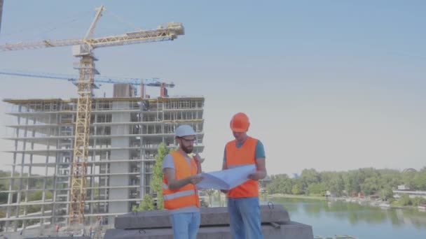 Ingenieros frente a un edificio de varios pisos. Dos ingenieros en un sitio de construcción están mirando el dibujo. Dos constructores en el fondo de la casa — Vídeos de Stock